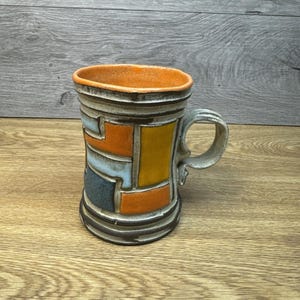 May include: A handmade ceramic mug with a geometric design. The mug features a light gray handle and rim, with a vibrant orange interior. The exterior is adorned with colorful rectangular shapes in orange, yellow, blue, and gray.