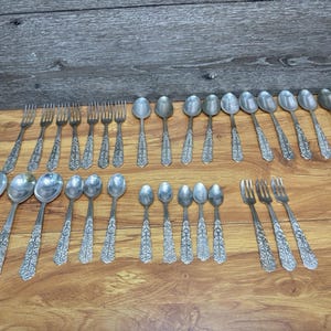 May include: A collection of silver-toned metal flatware featuring a decorative embossed pattern on the handles. The set includes various sizes of forks and spoons arranged in rows, showcasing a consistent floral-inspired design on each handle.