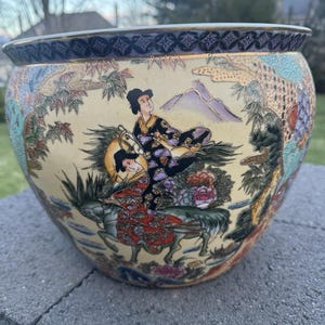 Könnte beinhalten: Eine dekorative Keramikschale mit breiter, runder Form. Die Schale hat einen cremefarbenen Hintergrund mit einer detaillierten Malerei im japanischen Stil mit Figuren, Bergen und floralen Elementen. Der Rand hat einen dunkelblauen und goldenen Musterbord.