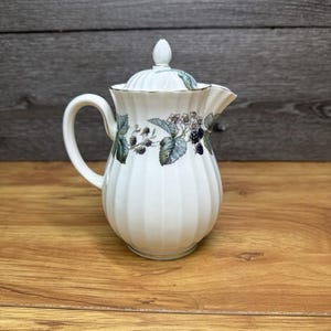 May include: A white porcelain creamer with a fluted, ribbed body and a matching lid. The piece features a delicate botanical pattern around the upper section, depicting green leaves, small white flowers, and dark purple blackberries.