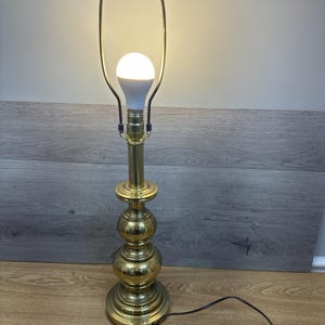 May include: A brass table lamp with a spherical base and a tall, slender body. The lamp has a white bulb and a dark brown power cord. The lamp's shade holder is a gold colour.