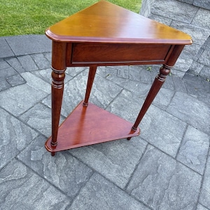 May include: A triangular wooden corner table with a smooth, polished reddish-brown finish. The table features a flat top surface, a lower triangular shelf, and three turned legs that provide structural support for the tiered design.