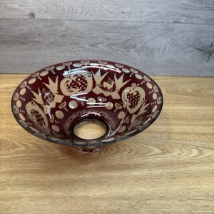 May include: A burgundy-colored glass bowl with a decorative etched design. The bowl features a wide, shallow shape with a central hole and is adorned with intricate patterns of stylized birds, fruits, and swirls. The etched details are in a lighter, contrasting color.