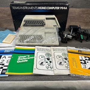 May include: A vintage Texas Instruments Home Computer 99/4A with keyboard, manuals, and accessories. The box is black with white text. The manuals include a Beginner's BASIC guide and a User's Reference Guide.