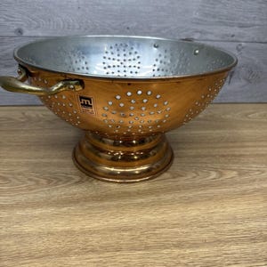 May include: A copper-coloured colander with a silver interior and small drainage holes. It has two brass handles and a pedestal base. The colander has a label that reads "Made in Portugal".