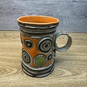 May include: A handmade ceramic mug with an orange interior and exterior. The mug features a gray handle and is decorated with orange and green circles. The mug has a rustic, textured appearance.