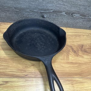 May include: A black cast iron skillet with a long handle featuring a hole for hanging. The skillet has a rounded shape with a slightly raised edge. The surface of the skillet appears well-seasoned, indicating it's ready for cooking.