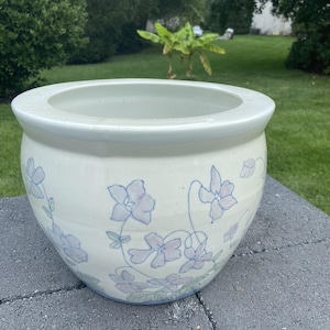 May include: A round, off-white ceramic planter with a wide rim. The planter is decorated with hand-painted, light purple floral designs and green stems. The planter is sitting on a gray brick surface, with a green lawn and trees in the background.
