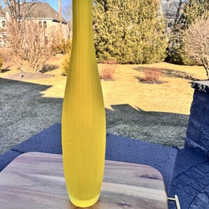 Puede incluir: Un jarrón alto de vidrio amarillo con un diseño delgado y curvo. El jarrón tiene una superficie texturizada y una parte superior asimétrica. Se exhibe sobre una tabla de cortar de madera, lo que sugiere un artículo decorativo o de decoración del hogar.
