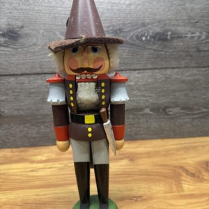 May include: A wooden nutcracker figurine in a soldier's uniform. The figurine has a brown hat, a mustache, and a stern expression. It wears a brown jacket with red and white accents, a black belt, and a silver knife.