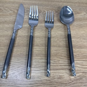 Könnte beinhalten: Ein Edelstahl-Besteckset mit einem Messer, zwei Gabeln und einem Löffel. Die Griffe haben eine dunkelgraue Oberfläche mit einem gedrehten Design. Das Besteck liegt auf einer hellbraunen Holzoberfläche.