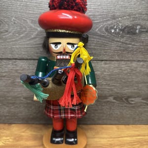 May include: A wooden nutcracker figure dressed as a Scottish bagpiper, wearing a red tam o' shanter with a red and black pom-pom, a green jacket, and a red plaid kilt. The figure holds a set of bagpipes with yellow, red, and blue tassels.