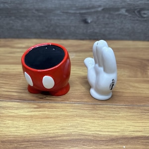 Puede incluir: Un salero de cerámica rojo y blanco con forma de cabeza de Mickey Mouse y un pimentero de cerámica blanco con forma de guante de Mickey Mouse. El salero tiene un interior negro y dos puntos blancos. El pimentero es una mano blanca con el logo de Disney.