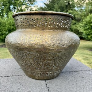 May include: Antique brass vase with intricate etched patterns and a decorative openwork rim. The vase has a wide, rounded body and a slightly flared base. The surface is aged with a warm, golden hue, showcasing detailed floral and geometric designs.