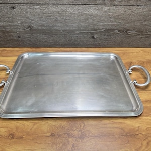 Puede incluir: Una bandeja de servicio rectangular de metal plateado con una superficie lisa y plana y un borde elevado. La bandeja cuenta con dos asas de metal curvadas en forma de semicírculo unidas a los lados izquierdo y derecho para facilitar el agarre.