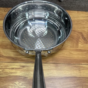May include: A stainless steel saucepan with a long handle. The interior features a perforated strainer. The pan sits on a wooden surface. The handle is angled towards the viewer.