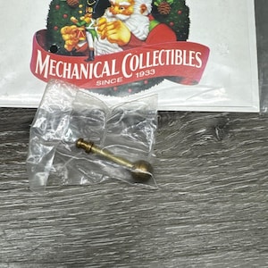 May include: A small brass-colored metal rod with a spherical end and a threaded shaft, enclosed in a clear plastic bag. Behind the bag is a white card featuring a graphic of Santa Claus, a nutcracker, and the text Mr. Christmas Mechanical Collectibles Since 1933.
