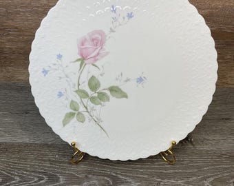 May include: White ceramic plate with a scalloped edge, featuring a pink rose and blue floral accents. The plate is supported by a gold-colored metal stand. The plate has a textured pattern around the edge.