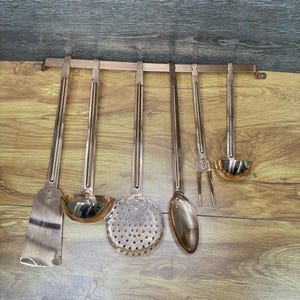 May include: A set of copper-colored kitchen utensils hanging on a wooden rack. The set includes a spatula, ladle, slotted spoon, spoon, fork, and a small ladle. The utensils are made of a shiny metal and are suspended against a wooden background.