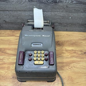 Peut inclure: Machine à additionner Remington Rand vintage de couleur grise avec un rouleau de papier. La machine possède un clavier numérique avec des touches jaunes et des boutons rouges. Le texte "Remington Rand" est visible.