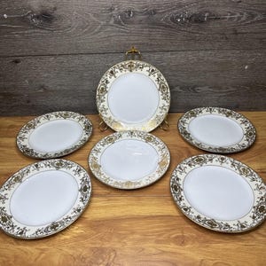 May include: A set of five white ceramic plates featuring a decorative gold filigree pattern along the rims. One plate is displayed upright on a gold metal stand, while the other four plates are arranged in a circular pattern on a flat surface.