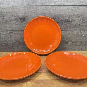 Könnte beinhalten: Drei runde Keramikteller in leuchtendem Orange. Jeder Teller weist eine Reihe konzentrischer, kreisförmiger Rillen auf, die entlang des äußeren Randes eingeprägt sind und einen strukturierten Abschluss zur glatten, glänzenden Oberfläche bilden.