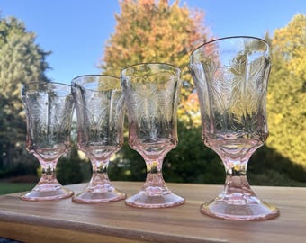 May include: Four pink-tinted vintage goblets with ornate floral and leaf patterns. The glasses have a flared shape and sit on a short stem and base. The goblets are arranged on a wooden surface, with a blurred background of trees and a blue sky.