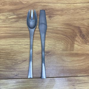 以下が含まれることがあります： 木製の表面に、3本歯のフォークとナイフが置かれています。フォークとナイフはどちらもグレーで、マットな仕上がりです。ナイフは平らで丸みを帯びた刃をしています。