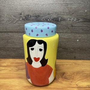 Op de afbeelding: Een gele keramische pot met een deksel. De pot heeft een geschilderd vrouwengezicht met zwart haar, een rood shirt en een rode lip. Het deksel is lichtblauw met paarse stippen. De pot staat op een houten oppervlak.