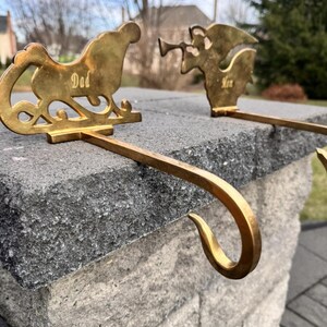 May include: Gold-colored Christmas stocking holders. One is shaped like a sleigh with the word "Dad", and the other is an angel with the word "Mom". The holders have a curved hook for hanging stockings.