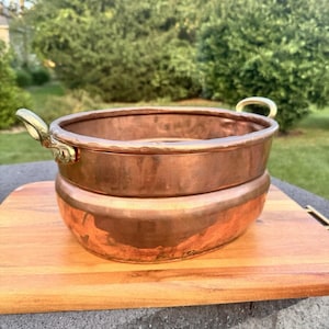 May include: Two copper bowls stacked on a wooden chopping board. The bowls have a shiny surface and brass handles. The background features green foliage and a stone surface.