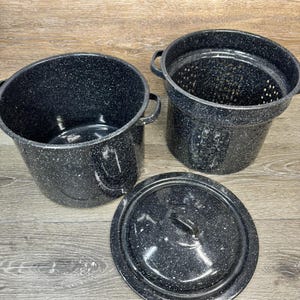 Può includere: Tre pentole in smalto nero maculato con manici e un coperchio abbinato. Le pentole sono di varie dimensioni, una con interno perforato. Lo smalto ha un classico design maculato, perfetto per cucinare o esporre.
