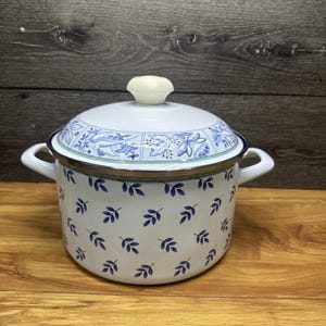 以下が含まれることがあります： 2つのサイドハンドルと一致する蓋が付いた白いエナメル鍋。鍋には小さな青い葉の繰り返し模様があります。蓋は青と白の花と鳥のモチーフで装飾されており、上部には無地の白いノブハンドルが付いています。