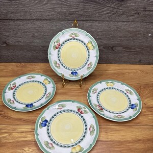 May include: Four decorative plates with a yellow centre and a white border. The plates feature a green and white scalloped edge, with a design of fruit and leaves. One plate is displayed on a gold stand.