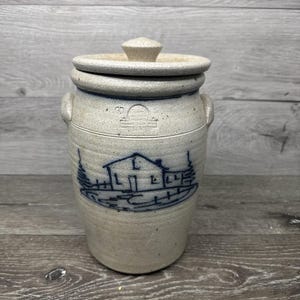 Può includere: Un barattolo in gres con coperchio, con un disegno di fattoria blu e bianco. Il barattolo ha due manici e un coperchio con un pomello. Il barattolo è cilindrico e poggia su una superficie di legno.