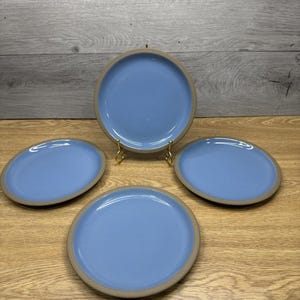 Pode incluir: Quatro pratos de cerâmica com um centro azul claro e uma borda bege estão dispostos sobre uma superfície de madeira. Os pratos são redondos e têm um design simples e moderno. Os pratos são um conjunto, perfeito para servir refeições.