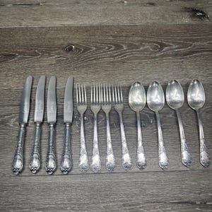 May include: A set of antique silverware, including knives, forks, and spoons, arranged on a wooden surface. The silverware has ornate handles and a polished, reflective finish, suggesting a vintage or heirloom quality.