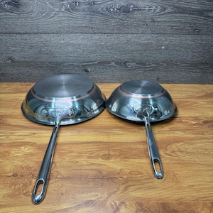 Puede incluir: Dos sartenes de acero inoxidable con mangos largos remachados. Las sartenes tienen una banda de color cobre alrededor de la base y un acabado pulido. Los mangos tienen un agujero para colgar. Las sartenes se exhiben sobre una superficie de madera.