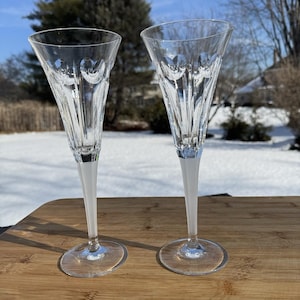 May include: Two clear crystal champagne flutes with etched heart designs. The flutes have a white stem and a clear, round base. The glasses are displayed on a wooden surface with a snowy background.