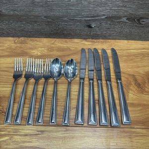 Op de afbeelding: Een set roestvrijstalen bestek, inclusief vorken, lepels en messen, gerangschikt op een houten oppervlak. Het bestek heeft een eenvoudig, elegant ontwerp met rechthoekige handvatten. De vorken, lepels en messen zijn allemaal gemaakt van glanzend, zilverkleurig metaal.