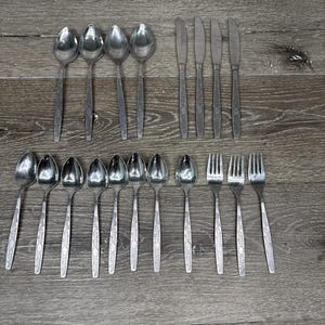 Peut inclure: Un ensemble de couverts en acier inoxydable, comprenant des cuillères, des couteaux et des fourchettes, disposés sur une surface en bois grise. Les couverts présentent un design simple et élégant avec un motif texturé sur les manches. Les cuillères, couteaux et fourchettes sont en bon état.