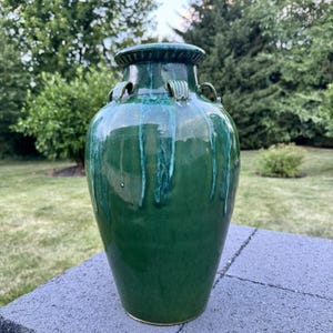 Op de afbeelding: Een glanzende groene keramische vaas met een brede rand en decoratieve handvatten. De vaas heeft een afgeronde body en een gladde afwerking met donkerdere groene druppels. De vaas staat tegen een achtergrond van groen gebladerte.