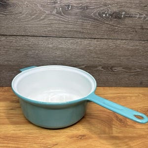 May include: A round, shallow enameled metal saucepan with a glossy turquoise exterior and a smooth white interior. The pan features a long, matching turquoise handle with a circular hole at the end and a small, short handle on the opposite side.
