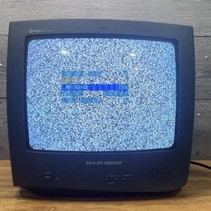 Puede incluir: Un televisor negro Philips Smart series con una pantalla que muestra estática y texto en pantalla. El televisor está sobre una superficie de madera con un fondo de madera. El televisor tiene un botón de encendido y un altavoz.