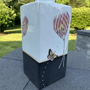May include: A square ceramic vase with a white upper section and a black lower section. The white section features a pink and yellow floral design and a butterfly. The vase is sitting on a gray surface.