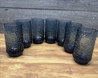 May include: A set of seven cylindrical drinking glasses in a dark charcoal gray hue. Each glass features a distinct textured, crinkled surface pattern that catches the light, with a smooth, slightly tapered base for stability and a clear rim.