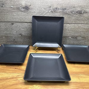 May include: A set of four matte black square plates with slightly raised edges. One plate is positioned upright on a gold-toned metal stand, while the other three plates are arranged flat in a triangular formation around the central upright plate.