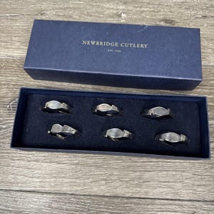 Puede incluir: Un juego de seis servilleteros plateados en una caja azul marino. La caja lleva la inscripción "NEWBRIDGE CUTLERY EST. 1934". Los servilleteros tienen un diseño ondulado y están dispuestos en una bandeja forrada de terciopelo azul oscuro.