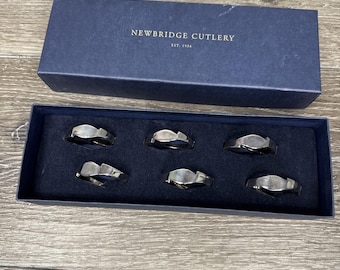 Op de afbeelding: Een set van zes zilverkleurige servetringen in een marineblauwe doos. De doos is voorzien van het opschrift "NEWBRIDGE CUTLERY EST. 1934". De servetringen hebben een golvend ontwerp en zijn gerangschikt in een donkerblauwe, met fluweel gevoerde lade.