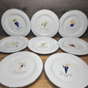 Könnte beinhalten: Ein Set aus acht weißen Keramiktellern mit silbernem Rand, die jeweils eine einzigartige Illustration eines Rentiers zeigen. Die Teller sind mit den Namen Donner, Blitzen, Comet, Cupid, Prancer, Vixen, Dancer und Dasher in Schwarz beschriftet.